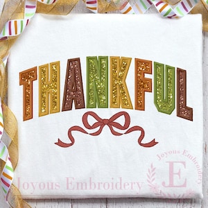 May include: Embroidered design of the word "THANKFUL" in a rainbow of autumn colours with a red bow underneath. The design is on a white fabric background.