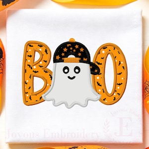 May include: A white fabric square featuring an embroidered design of the word BOO in orange with a black bat pattern. A white ghost wearing a black baseball cap with orange stars is positioned in the centre, replacing the letter O.