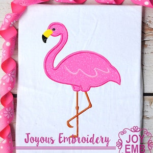 May include: Pink flamingo embroidery design with a yellow beak and legs. The design is on a white fabric background. Joyous Embroidery is written below the design.