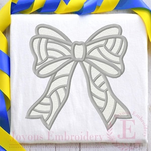 May include: A white embroidered bow with a detailed patterned design. The bow is on a white fabric background.