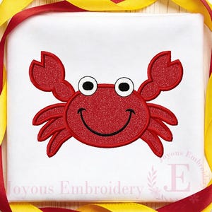 May include: A red crab design with a smiling face, black eyes, and claws, embroidered on a white background. The design is surrounded by red and yellow ribbons. The text "Joyous Embroidery" is at the bottom.