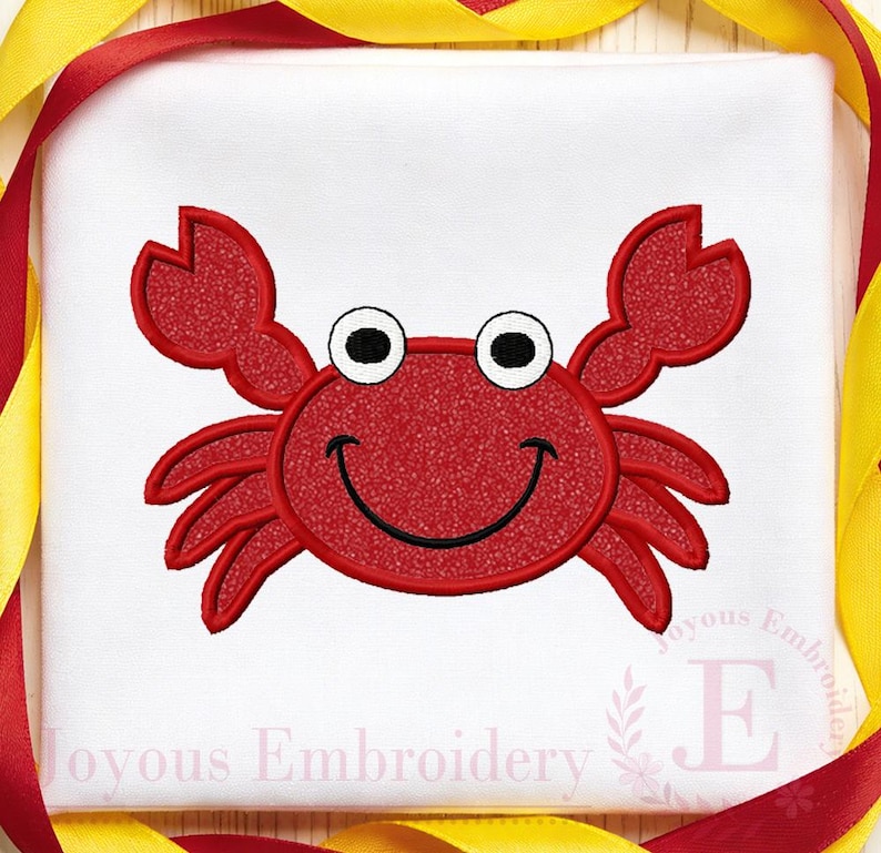 May include: A red crab design with a smiling face, black eyes, and claws, embroidered on a white background. The design is surrounded by red and yellow ribbons. The text "Joyous Embroidery" is at the bottom.