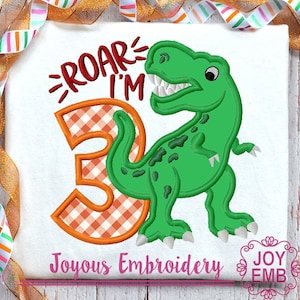 Peut inclure: Broderie représentant un dinosaure vert avec un chiffre 3 en vichy orange et blanc. Le texte "Roar I'm" est au-dessus du dinosaure.