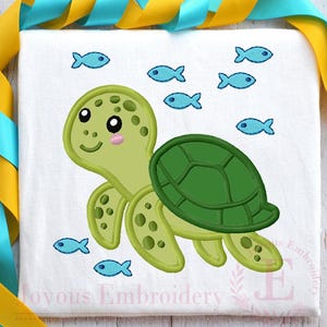 Op de afbeelding: Een wit T-shirt met een geborduurde cartoon schildpad. De schildpad heeft een groene schild en een lichtgroen lichaam, met een lachend gezicht. Blauwe vissen omringen de schildpad. De tekst "Joyous Embroidery" staat onderaan.