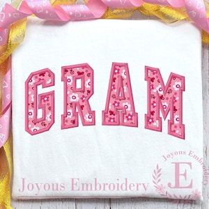 May include: White t-shirt with the word "GRAM" in pink, floral-patterned letters. The letters have a pink outline and are in a collegiate style. The text "Joyous Embroidery" is at the bottom, with a floral "E" logo.