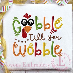May include: White t-shirt with the embroidered phrase "Gobble Till You Wobble" in colorful letters. The design incorporates a turkey face into the word "Gobble". The shirt is decorated with a gold ribbon.
