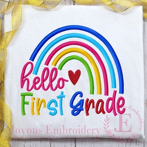 May include: White t-shirt with a colorful rainbow design. The rainbow arches over the words "hello First Grade" in bright, playful lettering. A red heart is nestled within the rainbow. The words "Joyous Embroidery" are at the bottom.
