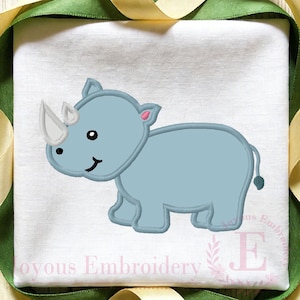 May include: A light blue embroidered rhinoceros design on a white fabric. The rhino has a white horn, black eyes, and a smiling mouth. The design is outlined in a darker blue, with pink accents on the ears. The fabric is framed with green and yellow ribbons.
