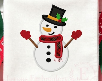 May include: A white fabric square featuring an embroidered snowman wearing a black top hat with a yellow band, green holly, and red berries. The snowman has an orange carrot nose, a red plaid scarf, and red mittens on brown stick arms.