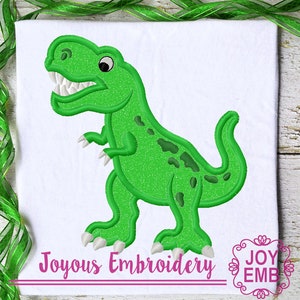 May include: Green embroidered dinosaur applique with white teeth and eyes. The dinosaur is standing on a white background with the text "Joyous Embroidery" and a logo that says "Joy Emb" in a decorative font.