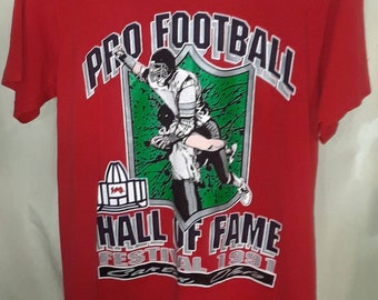 May include: Red t-shirt with a graphic of a football player in action. The graphic includes the text "Pro Football Hall of Fame Festival 1991".