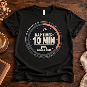 May include: A black short-sleeve t-shirt featuring a circular graphic resembling a timer. The text reads NAP TIMER: 10 MIN with a bed icon and the text ACTUAL: 2 HOURS below it. The design includes three Zs and a progress bar in peach.