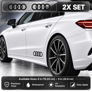 May include: A 2X set of black Audi logo vinyl decals applied to the side of a white car. The text 2X SET is visible at the top. Available sizes are 15.24 cm (6 inches) and 22.9 cm (9 inches). Features include premium vinyl, waterproof, and UV resistant.