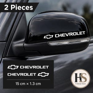 Könnte beinhalten: Ein Set aus zwei weißen Vinyl-Aufklebern mit dem Chevrolet-Logo und Schriftzug. Die Aufkleber sind auf einem schwarzen Fahrzeug-Außenspiegel angebracht und zusätzlich auf schwarzem Grund mit den Maßen 15 cm mal 1,3 cm abgebildet.