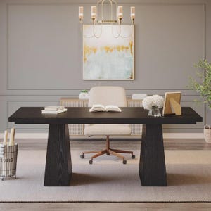 May include: A large rectangular black wooden desk with two wide trapezoidal legs. An off-white upholstered office chair sits behind the desk. On the desk surface are two stacked books, an open book, a white floral arrangement, and a framed picture.