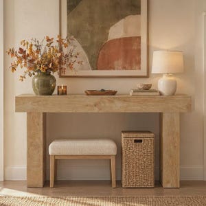 May include: A light-toned wooden console table with a thick rectangular top and sturdy legs. Underneath sits a beige upholstered bench and a tall woven wicker hamper. On the table are a green vase with autumn foliage, a candle, and a lamp.
