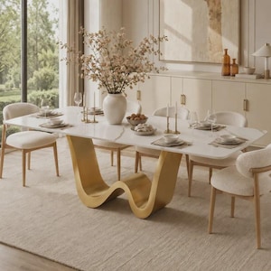 Op de afbeelding: Een eettafel met een wit rechthoekig blad en een goudkleurig, golfvormig onderstel. De tafel is gedekt met witte borden, glazen en gouden kandelaars. Een grote witte vaas gevuld met bloeiende takken staat in het midden van de tafel.