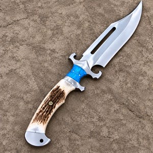 May include: A fixed-blade knife featuring a polished steel blade with a long cutout, a bright blue textured handle section, and a curved handle grip made of brown and cream-coloured antler material with a small brass pin detail at the centre.