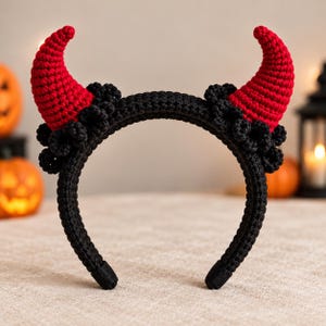 Pode incluir: Uma tiara de crochê preta com dois chifres vermelhos proeminentes em forma de cone voltados para cima. A base de cada chifre é cercada por um grupo de pequenas formas pretas de crochê que lembram flores, criando um contraste texturizado.