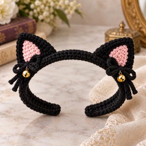 May include: A black crochet headband featuring two cat ears with light pink centers. Each ear is accented with a small black bow and a dangling gold-toned metal bell. The headband is constructed from thick black yarn with a visible textured stitch pattern.