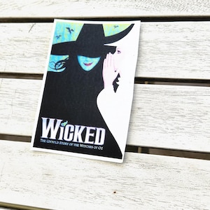 May include: A rectangular sticker featuring the Wicked musical logo with a black silhouette of a witch in a pointed hat and a white silhouette of a person whispering. The text reads Wicked The Untold Story of the Witches of Oz in white.