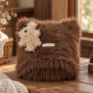 May include: A square brown faux fur pillow featuring a textured cream-colored teddy bear appliqué on the upper left corner and a small rectangular white fabric label with black text centered on the front surface of the plush material.