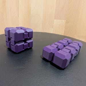 May include: Two purple fidget toys made of interconnected blocks. One toy is folded into a compact cube shape, while the other is unfolded into a flat, rectangular configuration. Both items feature a matte finish and a geometric, segmented design.