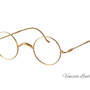 May include: Gold-toned metal spectacles featuring thin, perfectly circular frames and slender temples with a textured, coiled design. The bridge is curved and minimalist, connecting the two round rims. The text Venezia Lente is visible.
