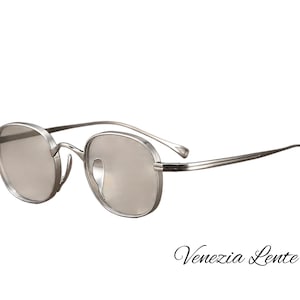 Puede incluir: Gafas de metal plateado con monturas redondas y lentes ligeramente tintadas. Las varillas metálicas delgadas tienen extremos curvos. El texto "Venezia Lente" está impreso en una fuente cursiva negra en la esquina inferior derecha.
