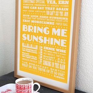May include: A yellow framed print with white text. The text is a word cloud of quotes from the British comedian Eric Morecambe. The text includes the phrases "Bring Me Sunshine", "Christmas Special", "Tea, Ern", "You Can Say That Again", "Now Look Here Sunshine", "Eric Morecambe", "Ernie Wise", "Get Out Of That Arsenal", "I'm Playing All The Right Notes", "The Play What I Wrote", "Just Watch It, That's All", "Short, Fat, Hairy Legs", "Singing In The Rain", "The Breakfast Sketch", "What Do You Think Of It So Far?" and "Rubbish".