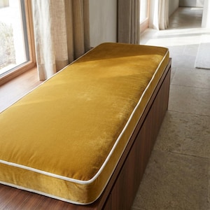 May include: A rectangular bench featuring a mustard yellow velvet cushion with white piping trim. The cushion sits atop a dark brown wooden base with a smooth, vertical grain finish, positioned horizontally in a bright, sunlit interior space.