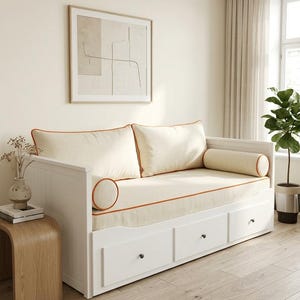 Peut inclure: Un lit de repos en bois blanc doté de trois tiroirs de rangement avec de petits boutons sombres. Le lit comprend un matelas crème, deux grands coussins de dossier rectangulaires et deux traversins cylindriques, ornés de passepoils orange.