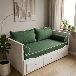 May include: A white daybed featuring a green fabric mattress and back cushions with cream-colored piping. The frame includes three lower storage drawers with small dark knobs and two cylindrical bolster pillows at each end of the seating area.