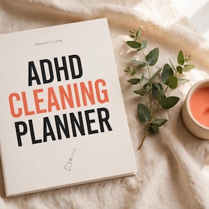 Könnte beinhalten: Ein weißer Planer mit schwarzem und korallenfarbenem Text, der PROJECT CLEAR ADHD CLEANING PLANNER lautet, sowie eine kleine Strichzeichnung eines Besens. Daneben liegen ein grüner Eukalyptuszweig und eine kleine runde Kerze mit korallenfarbenem Wachs.