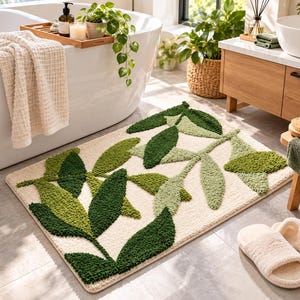 May include: A rectangular bath mat featuring a tufted, high-pile texture with a botanical pattern of leaves in varying shades of dark green, sage green, and olive green against a cream-colored background, creating a soft, plush surface.
