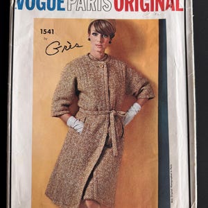 May include: A vintage Vogue Paris Original sewing pattern envelope, number 1541 by Grès. The cover features a photograph of a person wearing a textured, brown and white tweed coat with three-quarter sleeves, a tie waist, and a single button closure.