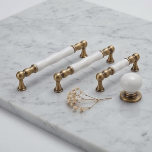 May include: Three cabinet pulls and one round cabinet knob featuring white ceramic centers with antique brass metal ends. The hardware pieces are arranged in a row, showcasing a smooth, curved design with metallic mounting bases.