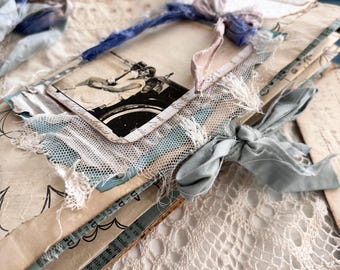 May include: A handmade junk journal featuring layered paper, vintage-style black and white photographs, frayed white netting, and blue silk ribbon ties. The cover includes printed text and decorative paper elements with a distressed, textured appearance.