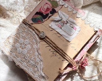 May include: A handmade journal featuring a cover with vintage-style floral paper, beige parchment, and white lace trim. It is wrapped in twine secured with a small metallic button and adorned with a fabric tag, pink dried flowers, and script text.
