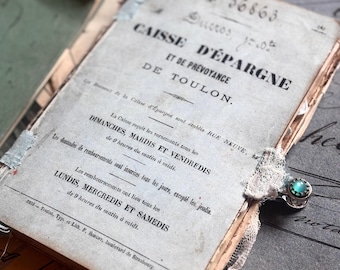 May include: An antique French savings bank booklet titled CAISSE D'ÉPARGNE ET DE PRÉVOYANCE DE TOULON with black printed text on a light gray cover, accented by a small silver ring featuring a round teal stone and white lace trim.
