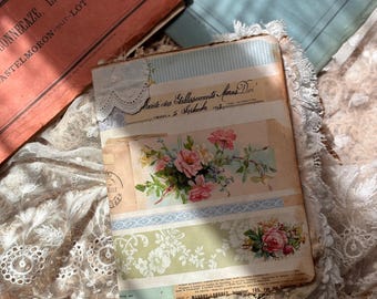 May include: A rectangular journal featuring a collage cover with floral illustrations of pink roses, green foliage, and vintage French text. The book is accented with lace trim along the edges and layered paper elements in shades of blue, green, and cream.