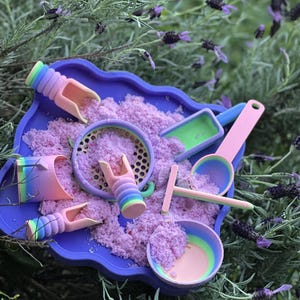 May include: A purple scalloped tray filled with pink sensory sand and a collection of rainbow-coloured plastic tools, including three small scoops with ribbed handles, a round sifter, a flat-bottomed shovel, a rake, and a small round bowl.