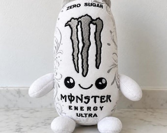 May include: A white plush toy shaped like a Monster Energy Ultra can, featuring a grey top, two arms, and two legs. The front displays the black Monster logo, the text ZERO SUGAR, MONSTER ENERGY ULTRA, and a cute face with black eyes.