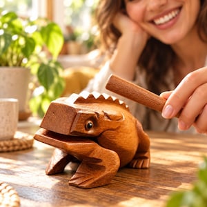May include: A carved wooden frog percussion instrument with a smooth, light brown finish, featuring a ridged back and a wide mouth. A matching wooden mallet is held near the frog, which is designed to be struck along its spine.