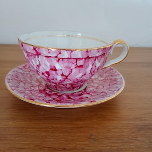 Puede incluir: Un juego de taza de té y platillo de porcelana con un vibrante patrón jaspeado en magenta y blanco. La taza tiene un asa curva y un borde dorado, mientras que el platillo a juego muestra el mismo diseño moteado en rosa y blanco con borde.