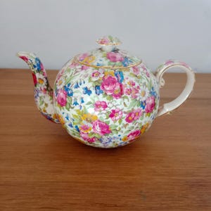 Può includere: Una teiera rotonda in porcellana caratterizzata da un vivace motivo floreale multicolore con rose rosa, campanule blu e margherite bianche. La teiera include un coperchio abbinato con un piccolo pomello, un beccuccio curvo e un manico dorato.