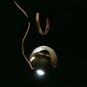 Op de afbeelding: Een ketting met hanger bestaande uit een doorschijnend, gebogen, gebroken wit harselement dat boven een gladde, goudkleurige metalen bol hangt. De bol reflecteert een heldere, geconcentreerde lichtbron tegen een donkere achtergrond.