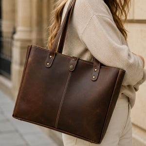 May include: A dark brown leather tote bag with a rectangular shape, featuring visible stitching, two shoulder straps, and a central snap button closure. The bag has brass-toned metal rivets securing the straps and the closure tab.