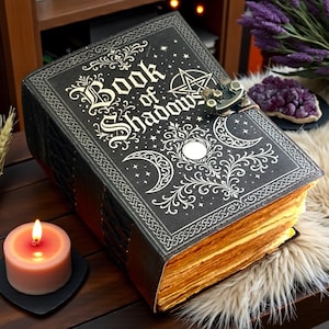 May include: A black hardcover book titled Book of Shadows with gold foil celestial designs including moons and stars. The book features a metal latch closure and thick, aged-looking paper pages. It is positioned on a surface with a lit pink candle.