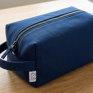 May include: A rectangular navy blue canvas toiletry bag featuring a top zipper closure, a side carry handle, and a small white fabric label with a black icon and the text "Tracey Digital Sewing" stitched onto the lower corner of the bag.